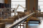 White robotic arms sorting boxes on a conveyor in an Amazon-style warehouse, official AWS Physical AI Toolchain announcement image